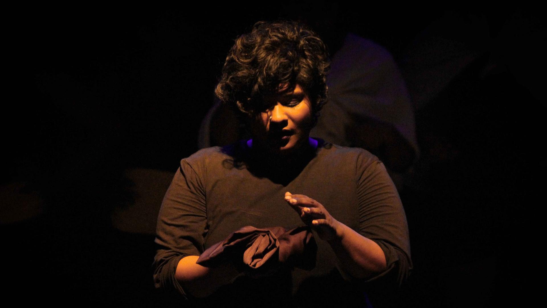 A performer wearing black is looking at a black satin cloth that she is holding in her right hand. Her left hand is hovering above the cloth.