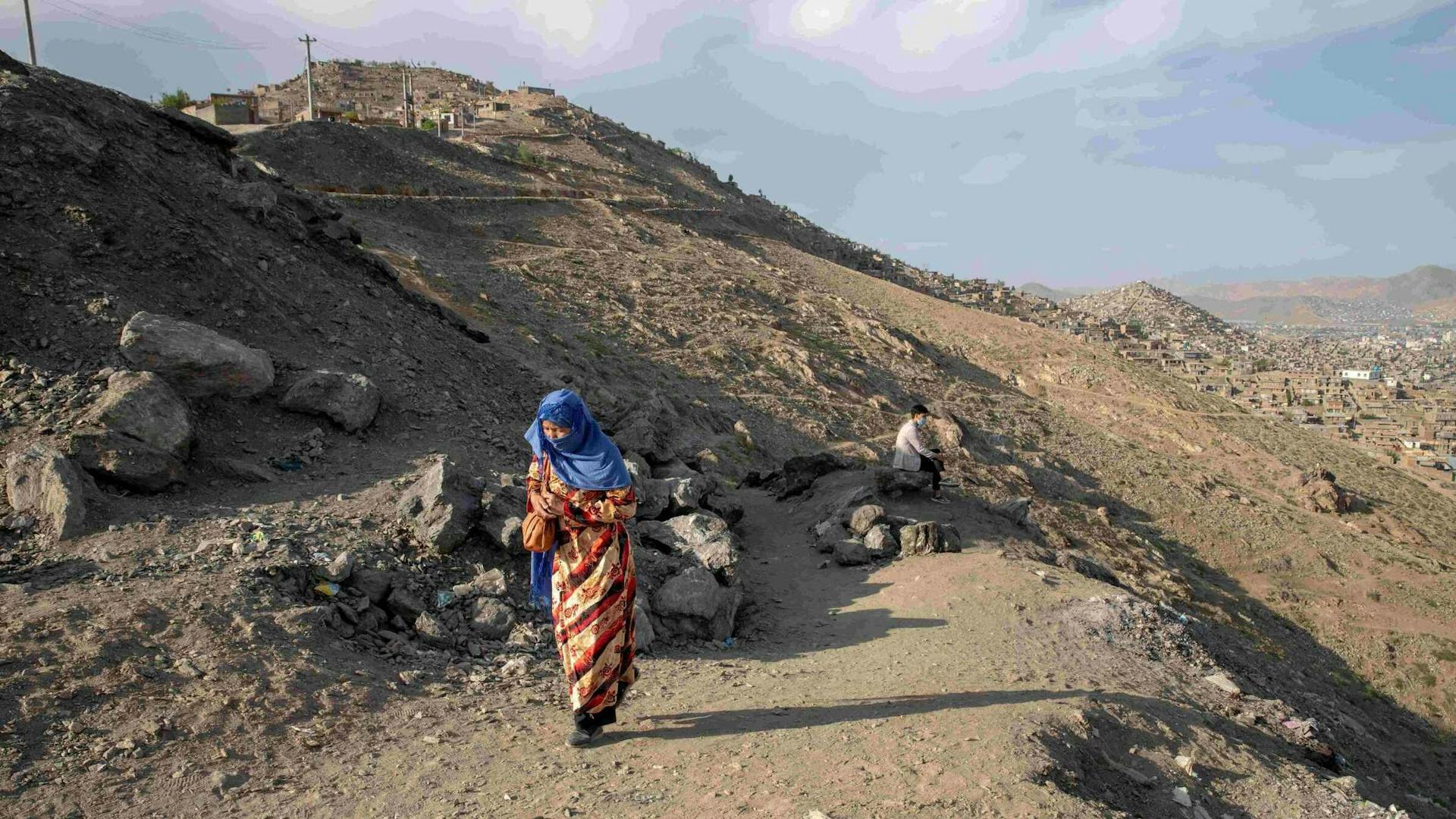 A woman in a blue headscarf walks alone down a dry mountain path.