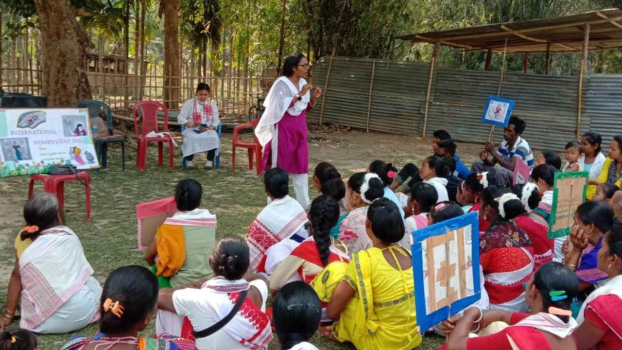 Pallabi Ghosh, dressed in pink and white, stands as she addresses a seated audience of rural community members during an outdoor training session. Some trainees hold paper signs with tape on the back of them.