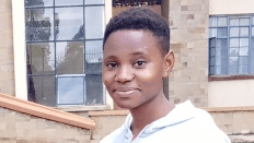 Faith Mwende, a woman with short dark hair wearing a white hoodie, smiles at the camera. Behind her are the windows of a building.