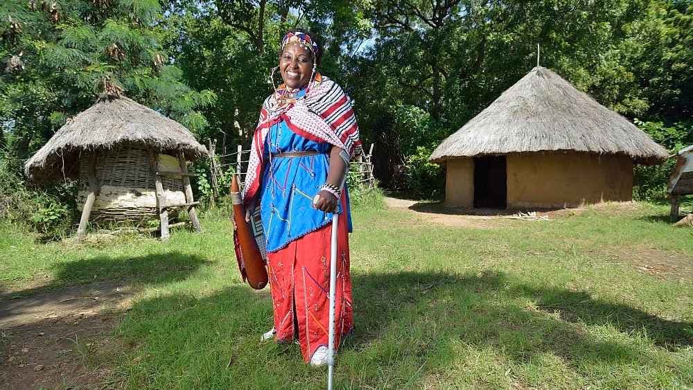 Jemimah Kutata stands smiling with an elbow crutch. She wears a blue and red dress with a colorful shawl and headpiece. The background is lush green with two thatched huts.