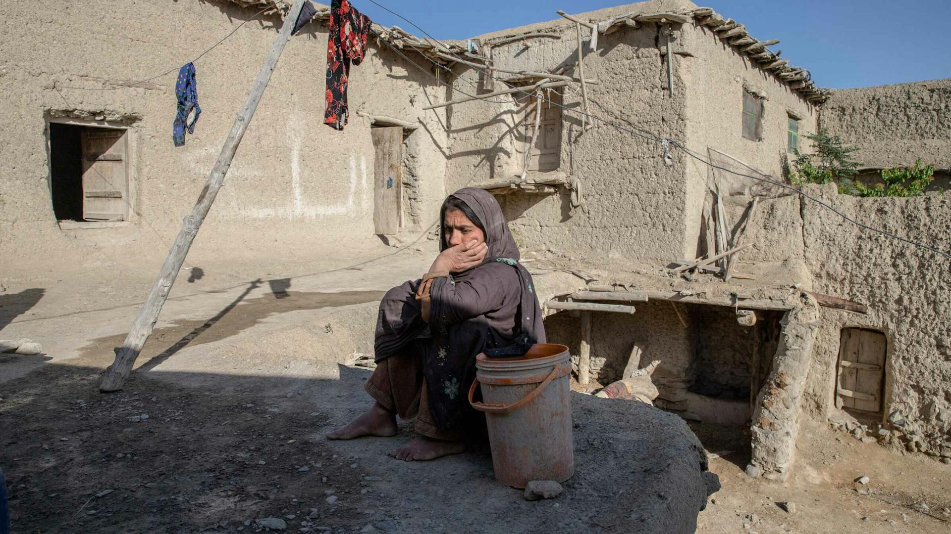 A woman in a hijab looks solemn as she sits alone in her dusty backyard.