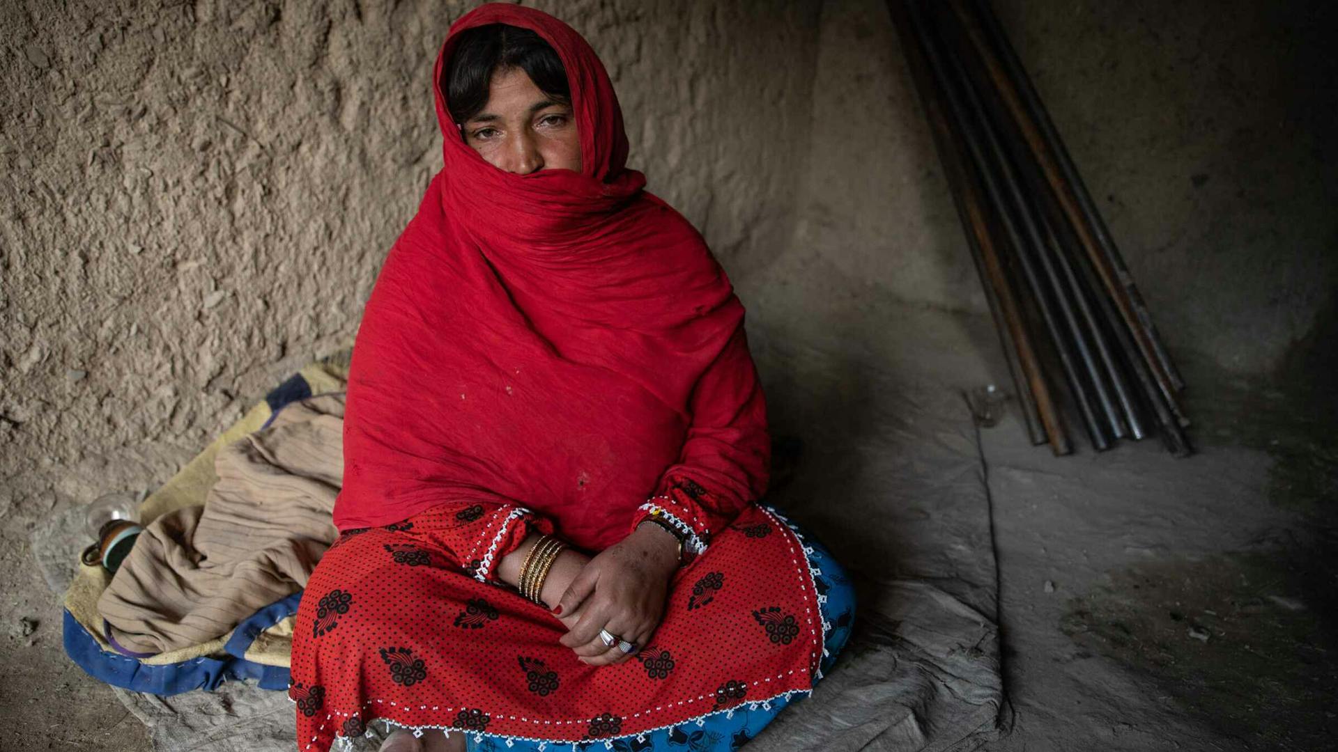 A young woman covered in a red veil sits on a blanky in a dim dirt hut.