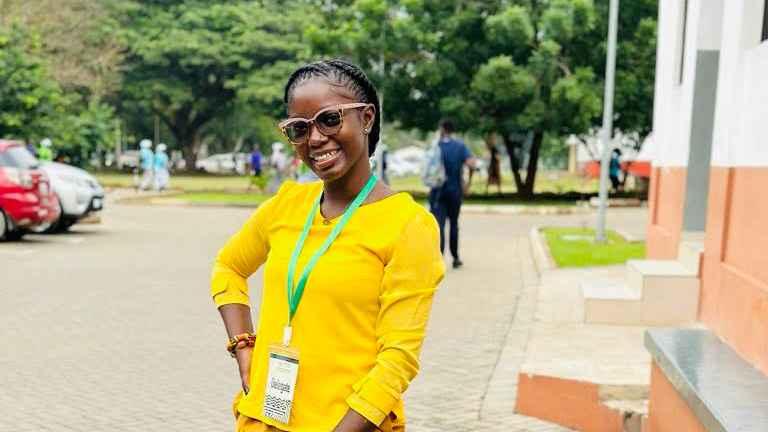 Fatmata Selina Kabba is photographed in a yellow top with dark braided hair, glasses, a lanyard, and a bracelet. Her hands are at her hips in front of a parking lots surrounded by trees.