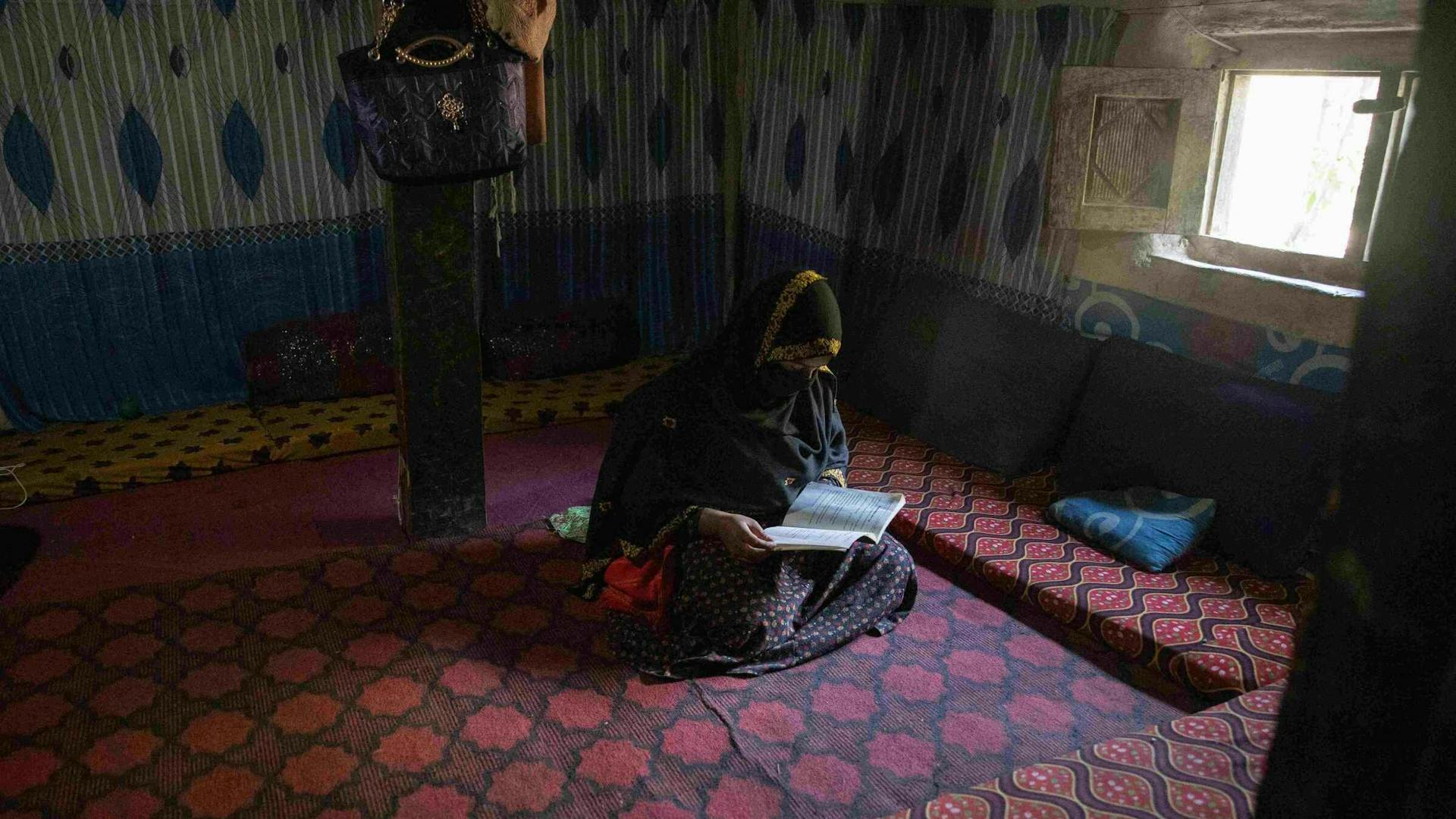 A veiled woman sits on a carpet and reads in a dimly-lit room.
