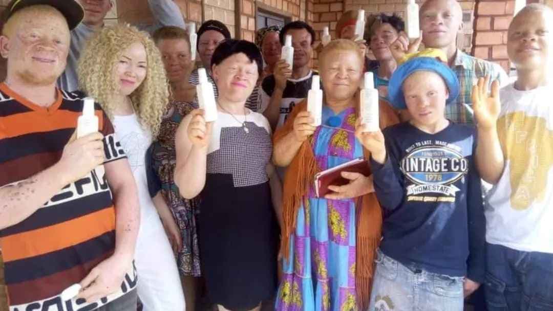 A group of 14 people with albinism stand and smile as they hold up white bottles containing products they made during a skin cancer workshop. 